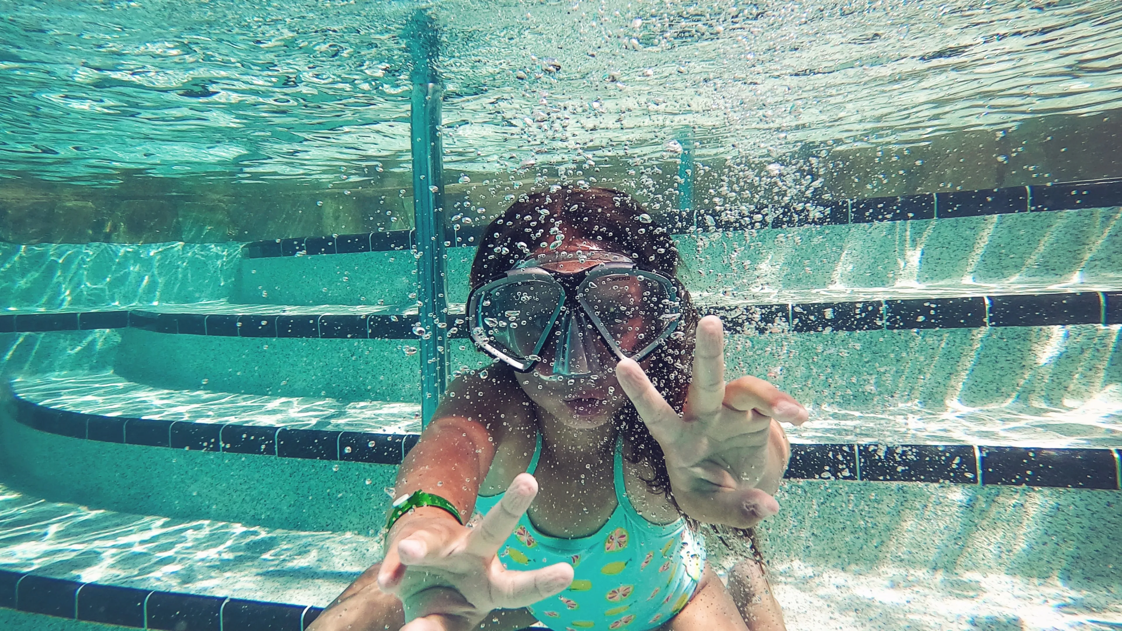 Child underwater in pool wearing snorkel mask, playfully forming peace signs with both hands.