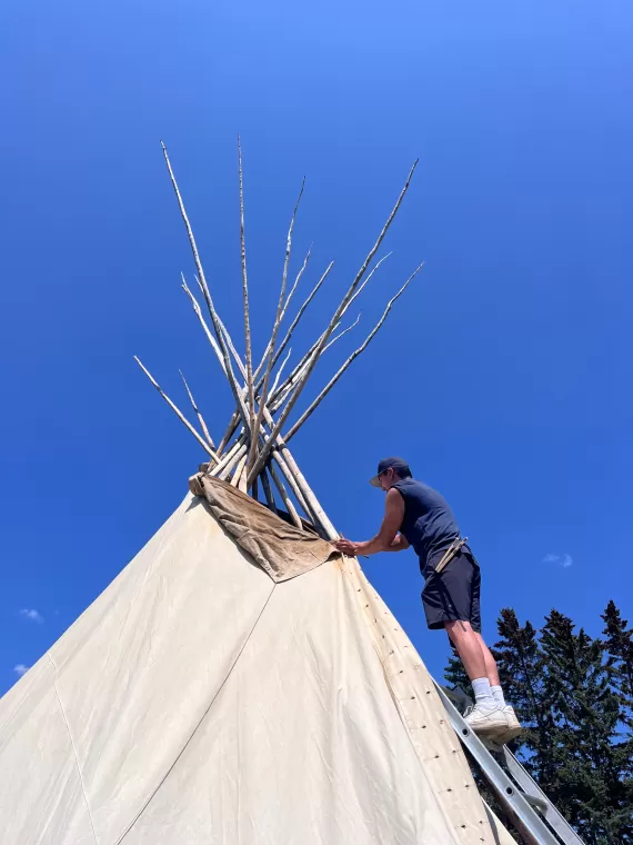 A person climbs a ladder to finishing pinning the canvas for a tipi at YMCA Calgary Camp Riveredge