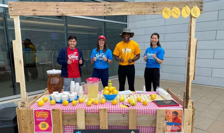 A group of 4 people stand in front of a wooden lemonade stand, smiling, in colourful t-shirts, to support the Camp It Forward campaign at YMCA Calgary.