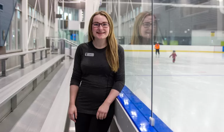 A young woman smiles while leaning against the glass of an ice rink at YMCA Calgary
