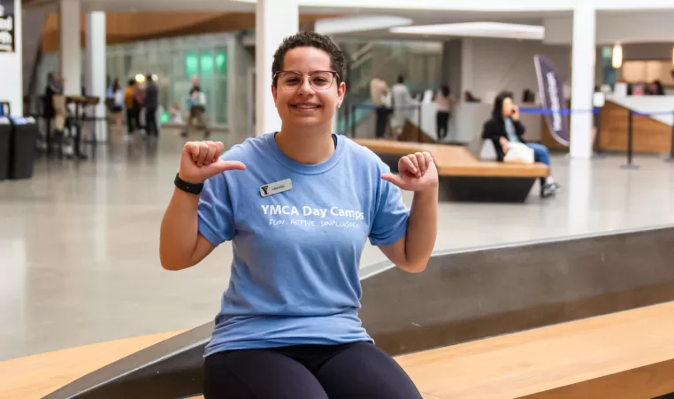 A young woman in a sky blue shirt that reads YMCA DAY CAMPS smiles and points to her shirt with her thumbs