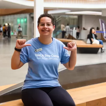 A young woman in a sky blue shirt that reads YMCA DAY CAMPS smiles and points to her shirt with her thumbs